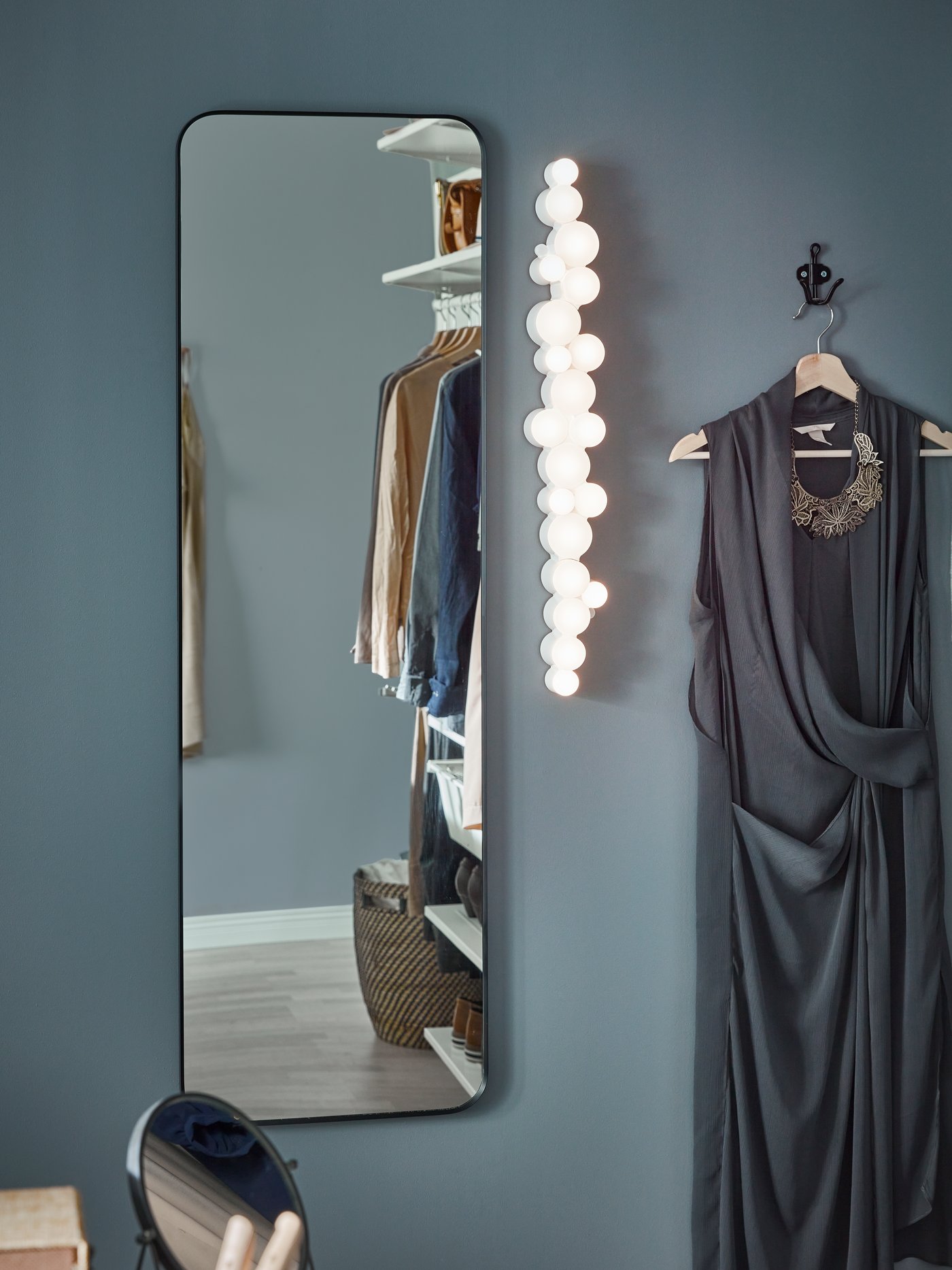 A LINDBYN mirror mounted on a dark blue wall. Beside it is an LED wall lamp and a grey evening dress on a hook.