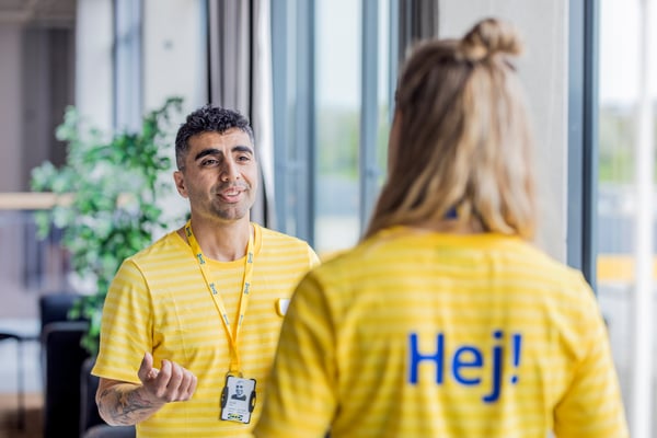 En ledertrainee i IKEA i en gul IKEA trøje står og taler med en person, der står med ryggen til i en gul IKEA trøje, hvor der står hej på ryggen