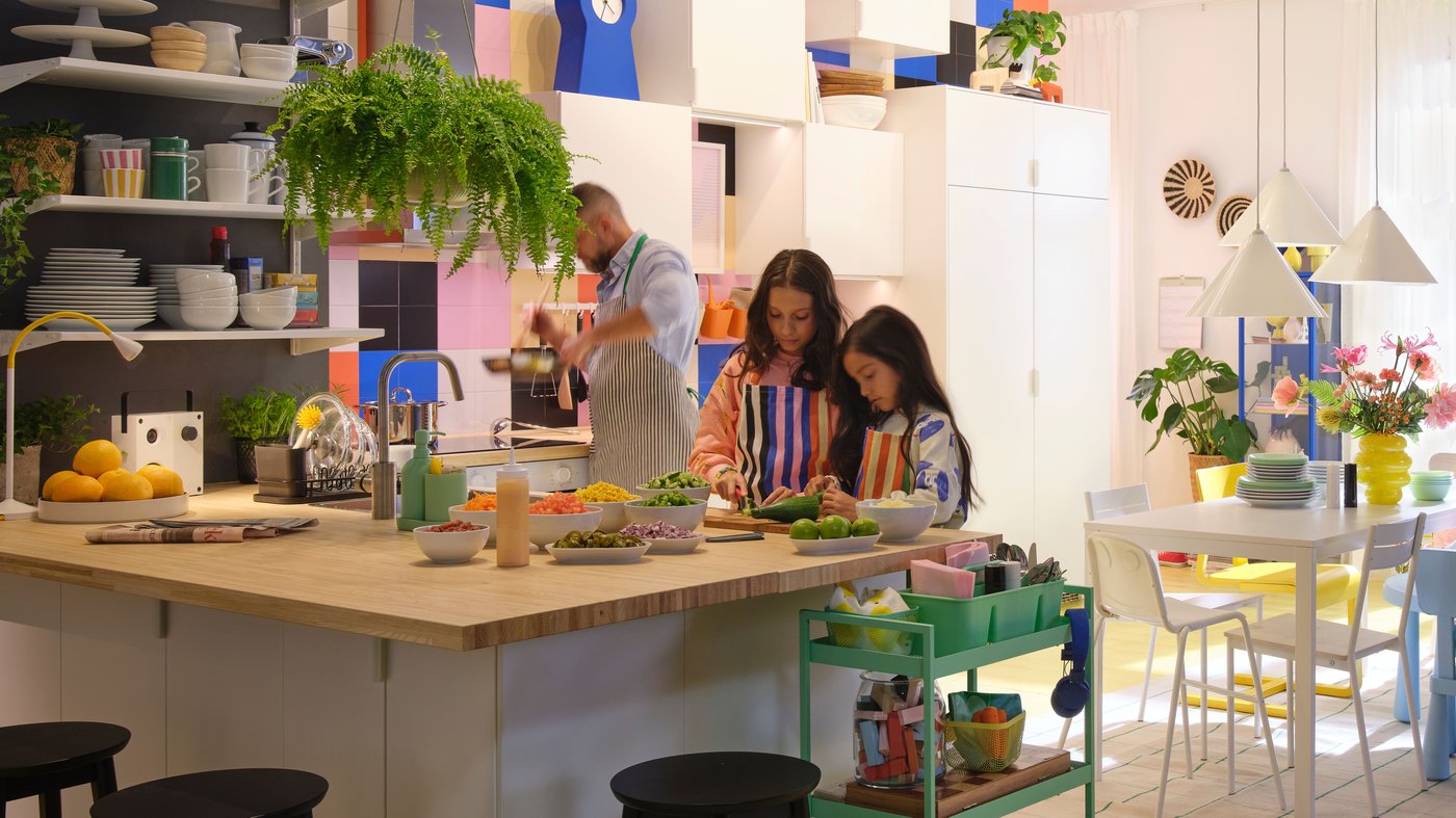 Dos niñas preparan alimentos sobre una encimera en acabado madera con varios platos y recipientes llenos de comida en una cocina de muebles blancos y azulejos de color