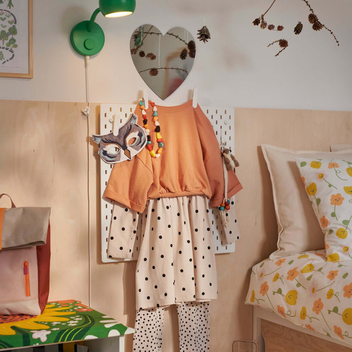 A beige and orange child’s outfit hangs on a SKÅDIS peg board topped by a HOLMÖARNA heart-shaped mirror.
