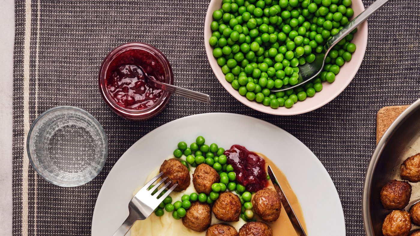A white plate has a portion of meatballs, mashed potatoes and green peas, with a jar of lingonberries and a glass next to it.