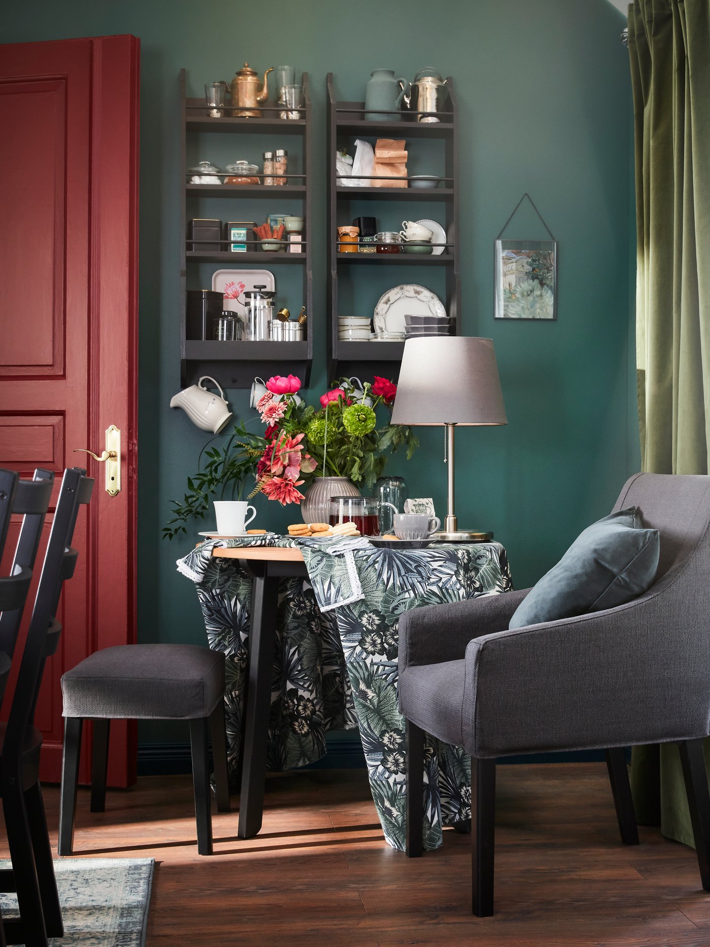 A corner with a black/dark grey chair with armrests, a table where tea is served and a window with white/green curtains.