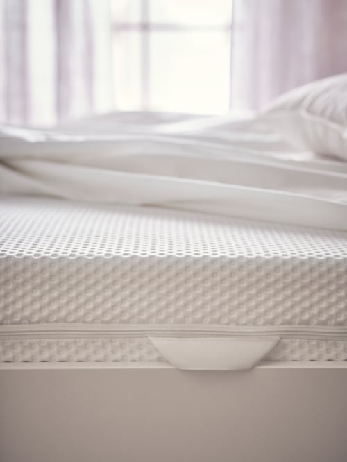 Prim-plan cu marginea parțială a  saltelei din spumă ÅBYGDA, cu mici denivelări. Pe saltea se află o lenjerie albă.