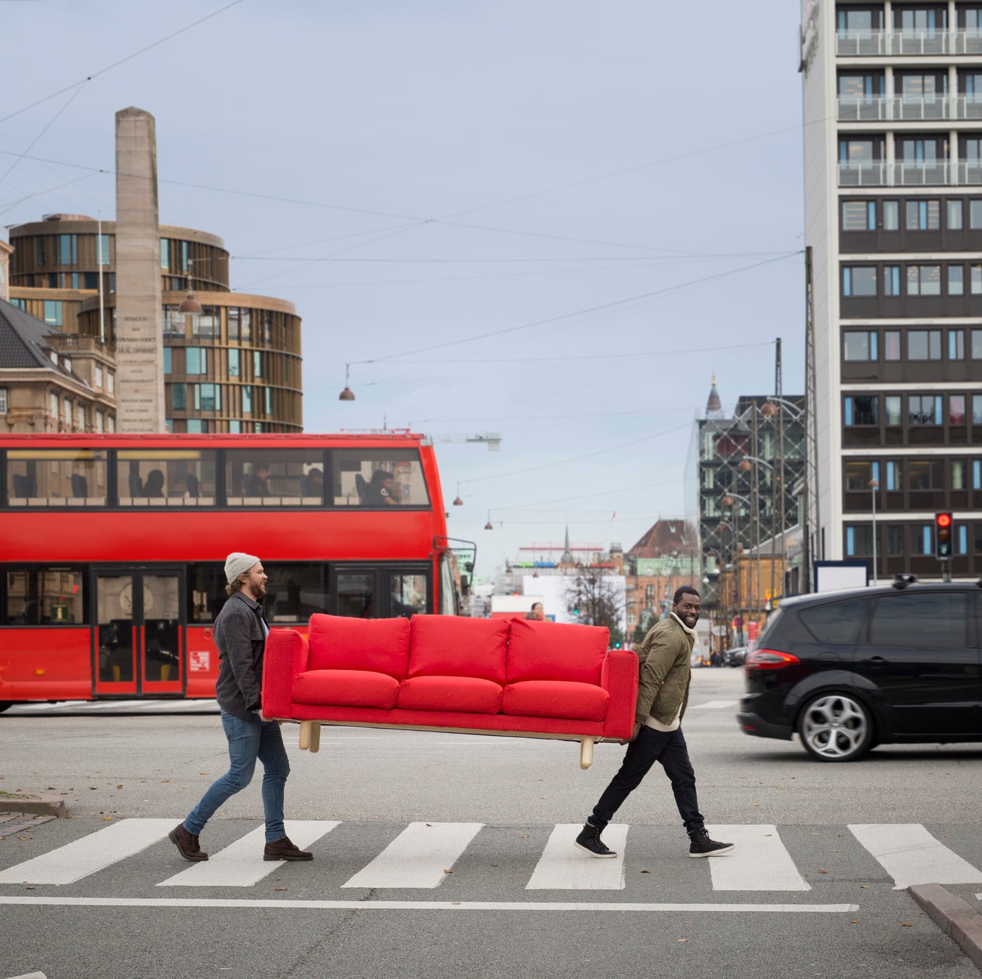 Two people carrying a red sofa in the middle of a street.