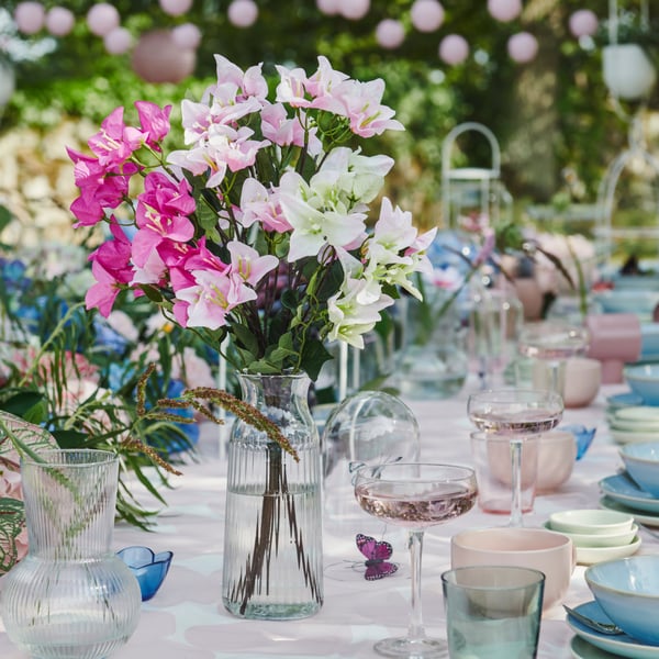 En kunstig SMYCKA buket med hvide og lyserøde blomster, står i en gennemsigtig VARDAGEN vase på et bord, der er dækket op til fest.