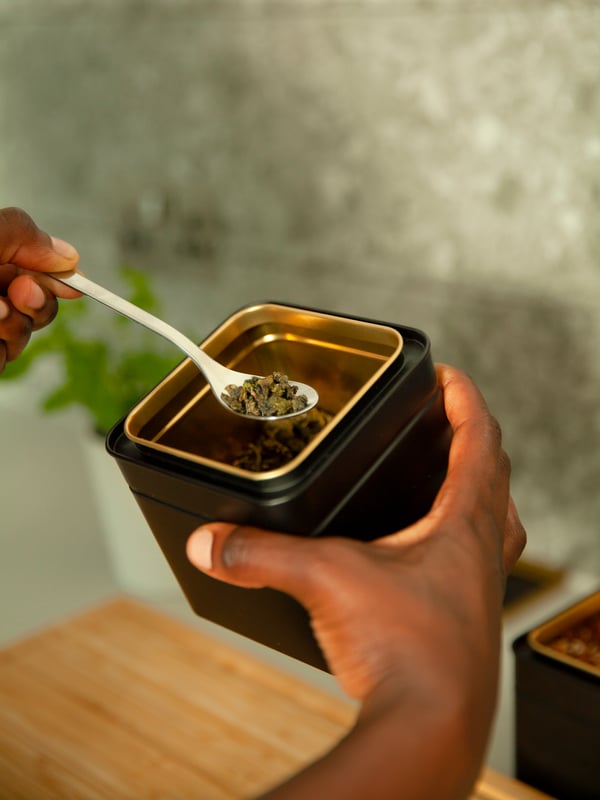 Someone is holding a white FÖRNUFT spoon above a BLOMNING teapot with plant food in it.