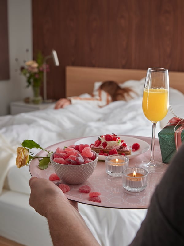 En person kommer gåetnde med en lyserrød bakke med et glas juide og en skål med hjerteformede vingummier til en kvinde der ligger og sover. 