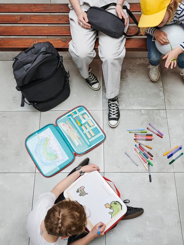 A group of small children are playing with a MALA portable drawing kit on a concrete floor, near a wooden bench. 