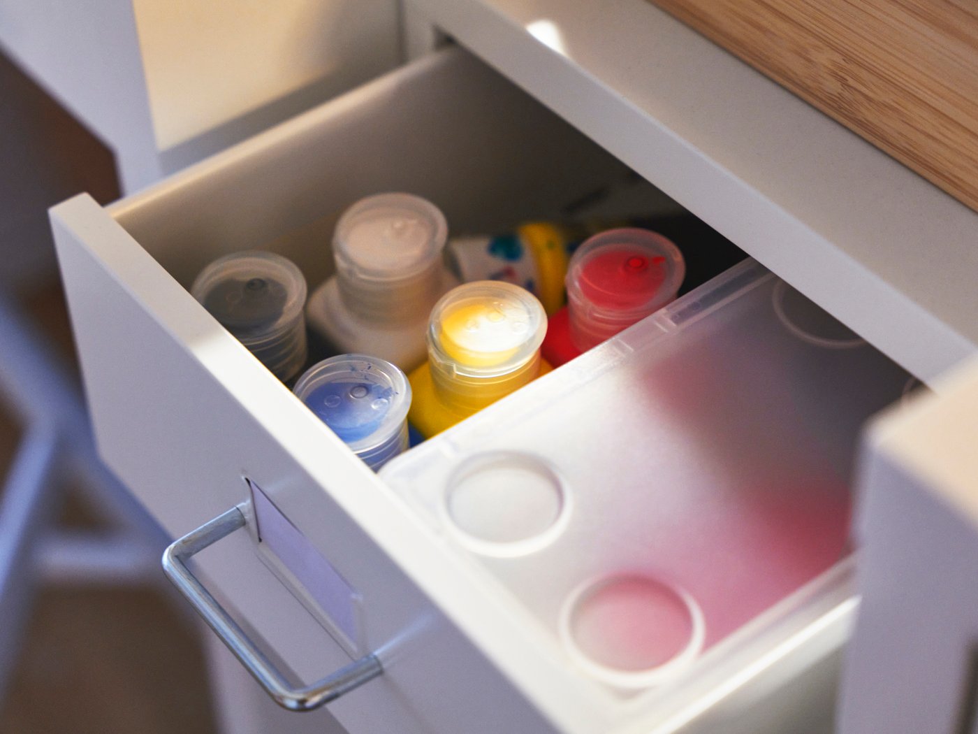 En öppen låda med små färgflaskor och en genomskinlig plastbricka placerad ovanpå, i en vit möbel.