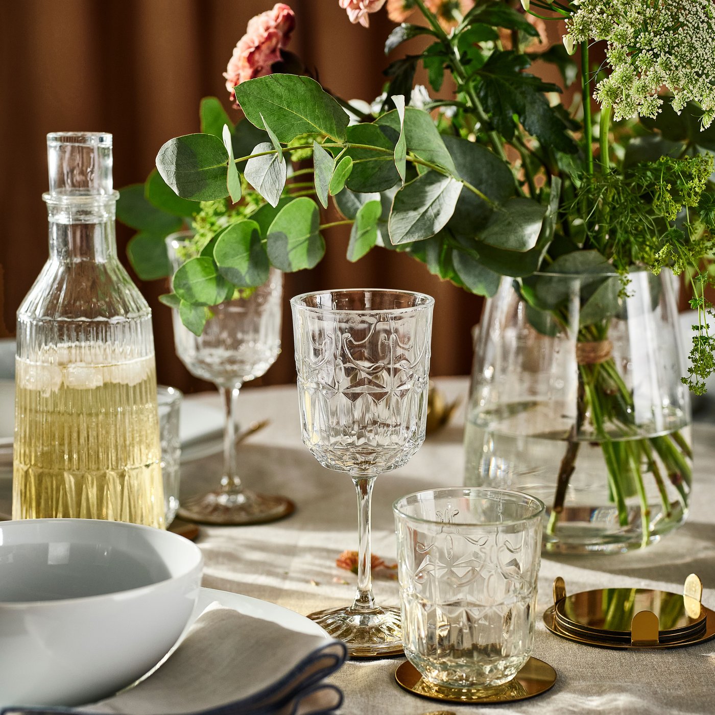 A SÄLLSKAPLIG glass carafe, glasses and wine glasses, plus flowers in a glass vase, on a table with a natural tablecloth.