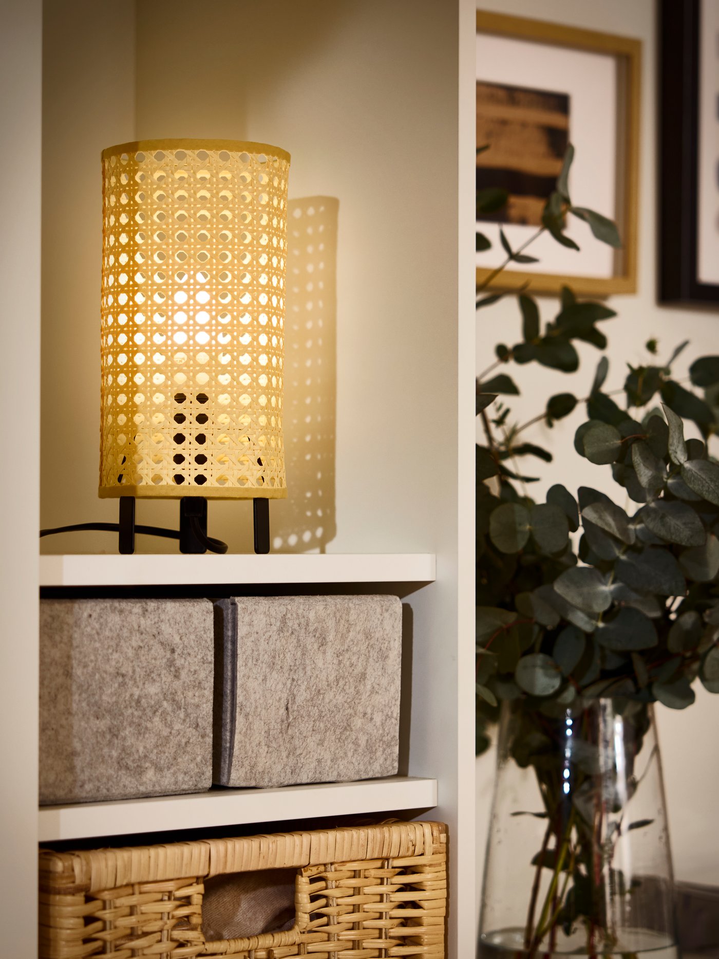 Část úložného dílu VIHALS, kde na policích stojí rozsvícená stolní lampa SAXHYTTAN, ratanový koš BRANÄS a úložné krabice.