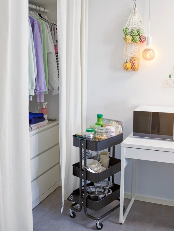 A RÅSKOG cart stands in the corner of a dorm room with cookware, plates, cups and food inside of it. Fruit hangs above.