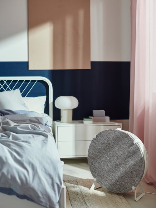 Dans une chambre à coucher, un purificateur d’air connecté STARKVIND au sol blanc avec une façade avant en tissu gris est sur le sol près du lit.