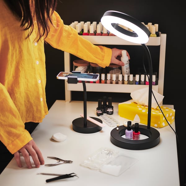 Una persona amb una brusa groga fa servir un anell lluminós LÅNESPELARE i un suport per a telèfon mentre es fa la manicura en una taula ben il·luminada.