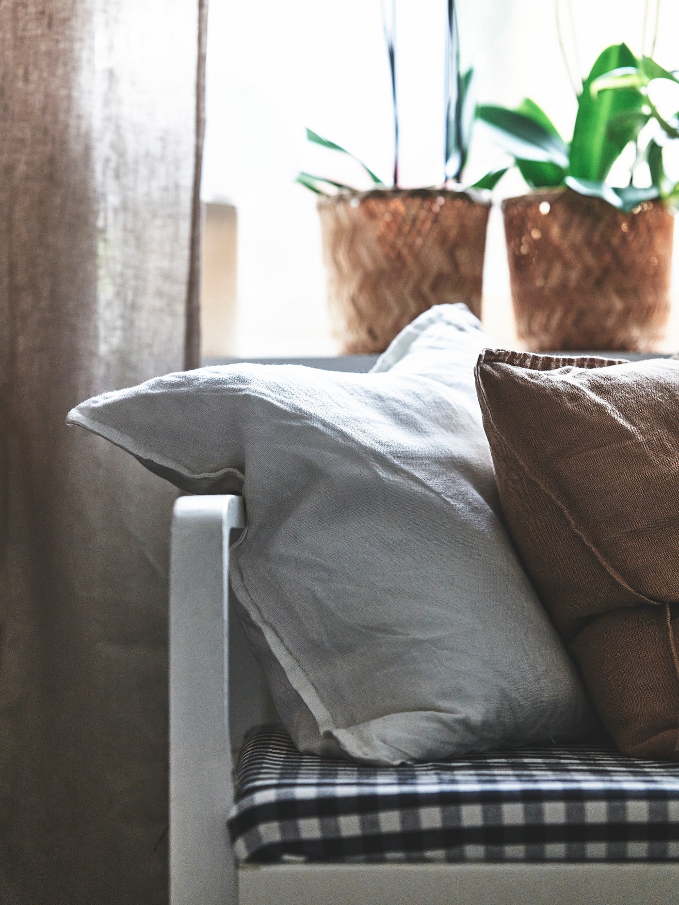 Närbild av en sittplats med två kuddar, en ljus och en brun, placerade på en bänk med mörkt rutigt sittunderlag. I bakgrunden står två krukväxter på en fönsterbräda.