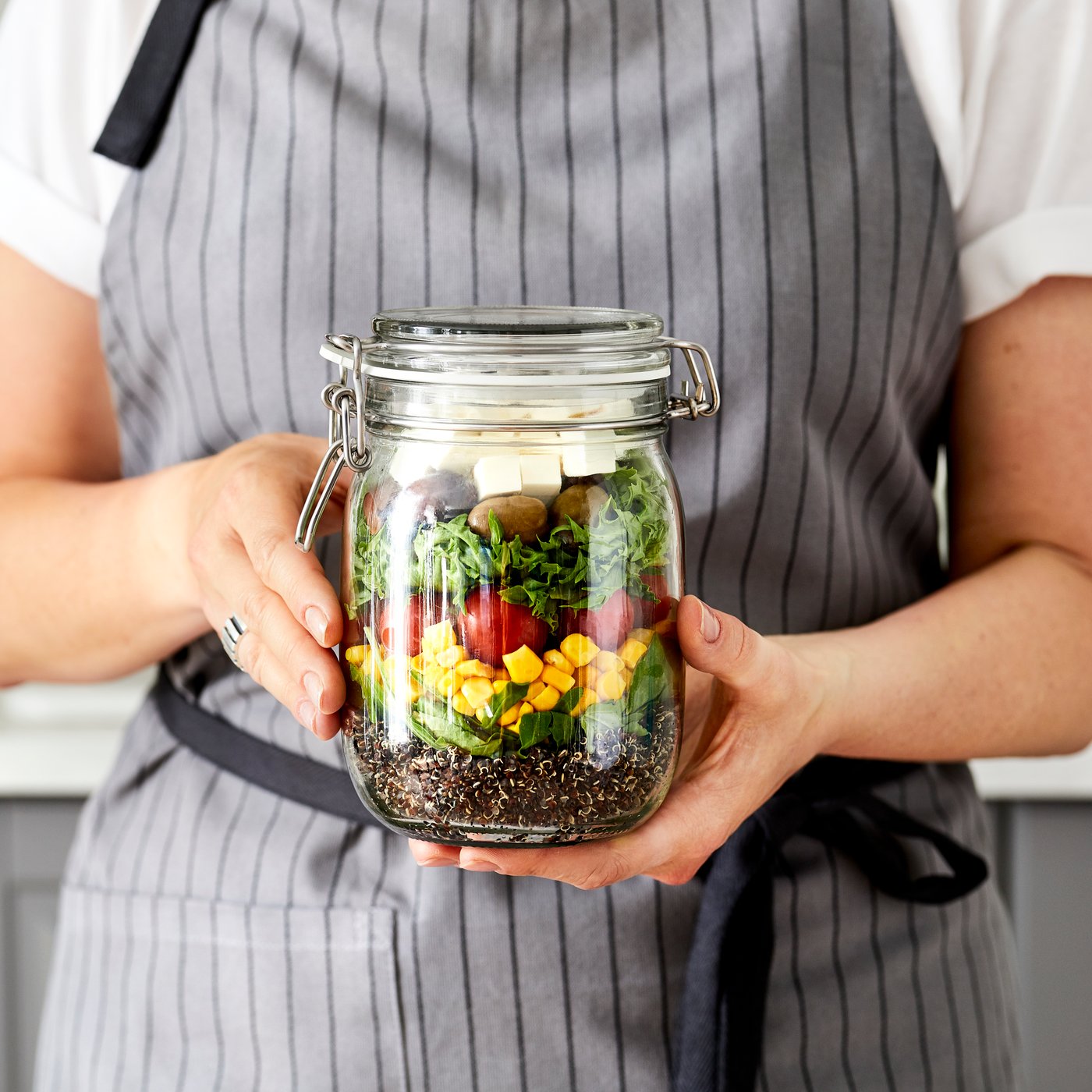 Woman in apron standing in kitchen, a loose two-hand grip holding up a tall glass jar filled with a layered, colourful salad.
