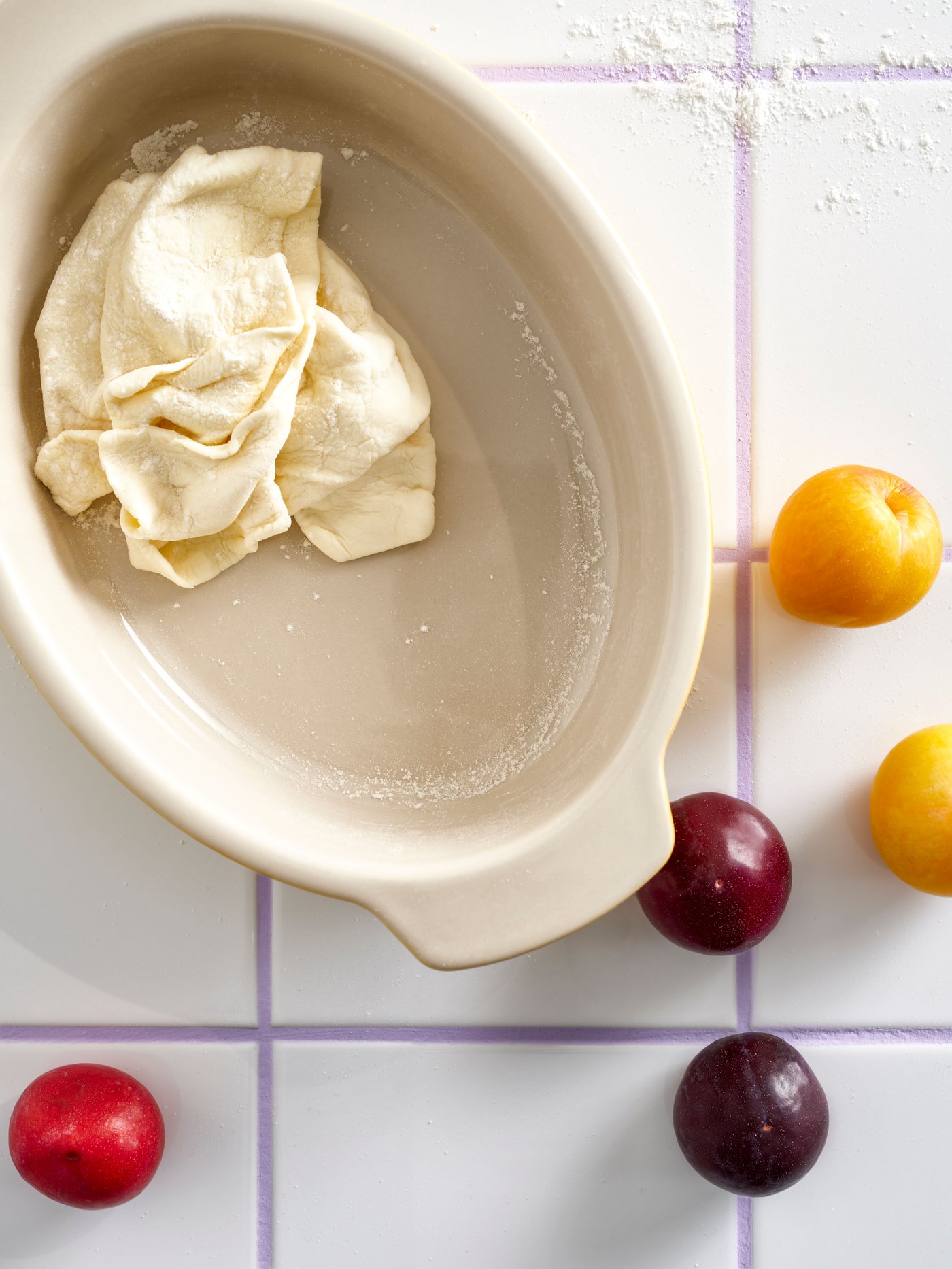 A POETISK oven dish seen from above with pastry dough in it and some plums and flour around it, on a white tiled surface.