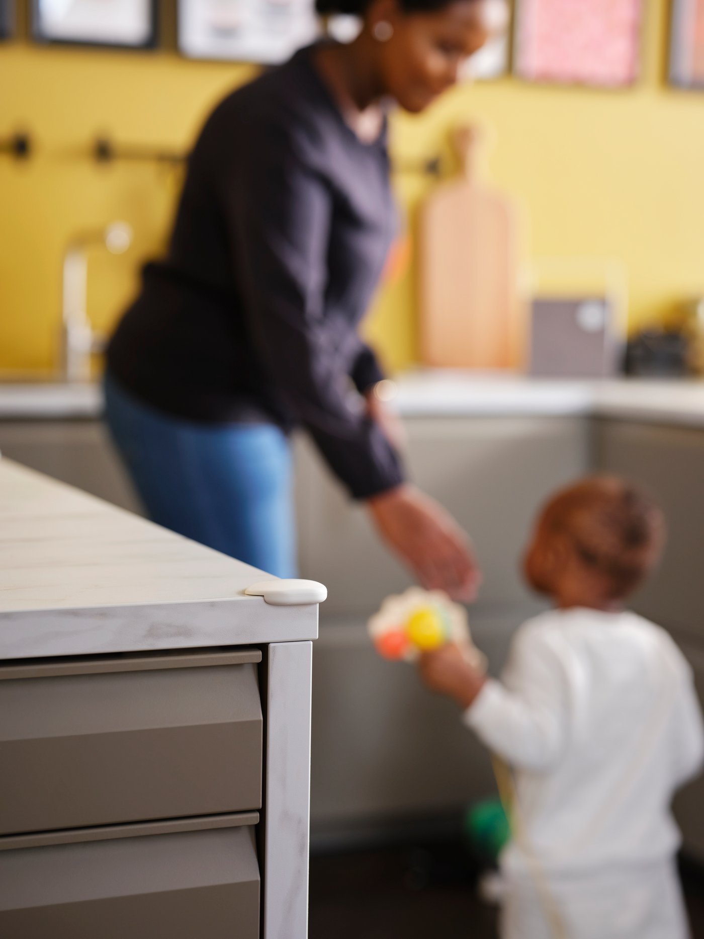 A woman and a toddler in a kitchen with a white UNDVIKA corner bumper on the corner of a white marble effect worktop.