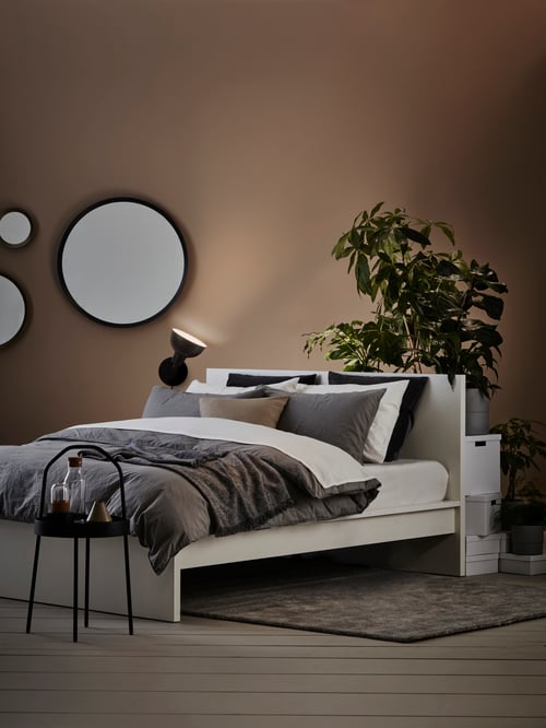 Bedroom with bed frame, duvet cover, pillowcases, side table, mirrors, uplighter, plants, pots, boxes, cushion covers, rug.
