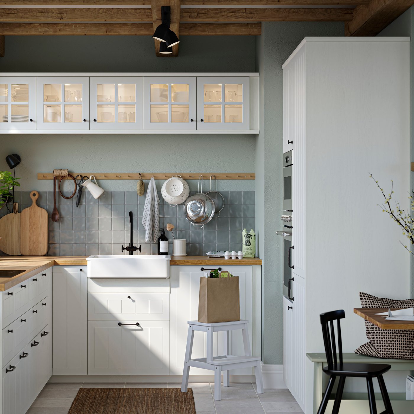 مطبخ بشكل حرف L وجداران بلون رمادي أخضر له واجهات STENSUND بيضاء وعلاقة خشبية مثبتة على الجدار بمقابض متعددة.