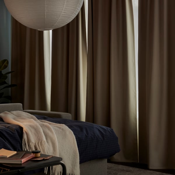 Beige KORGMOTT block-out curtains elegantly drape in a cosy living room, partially opened to reveal soft, natural light.