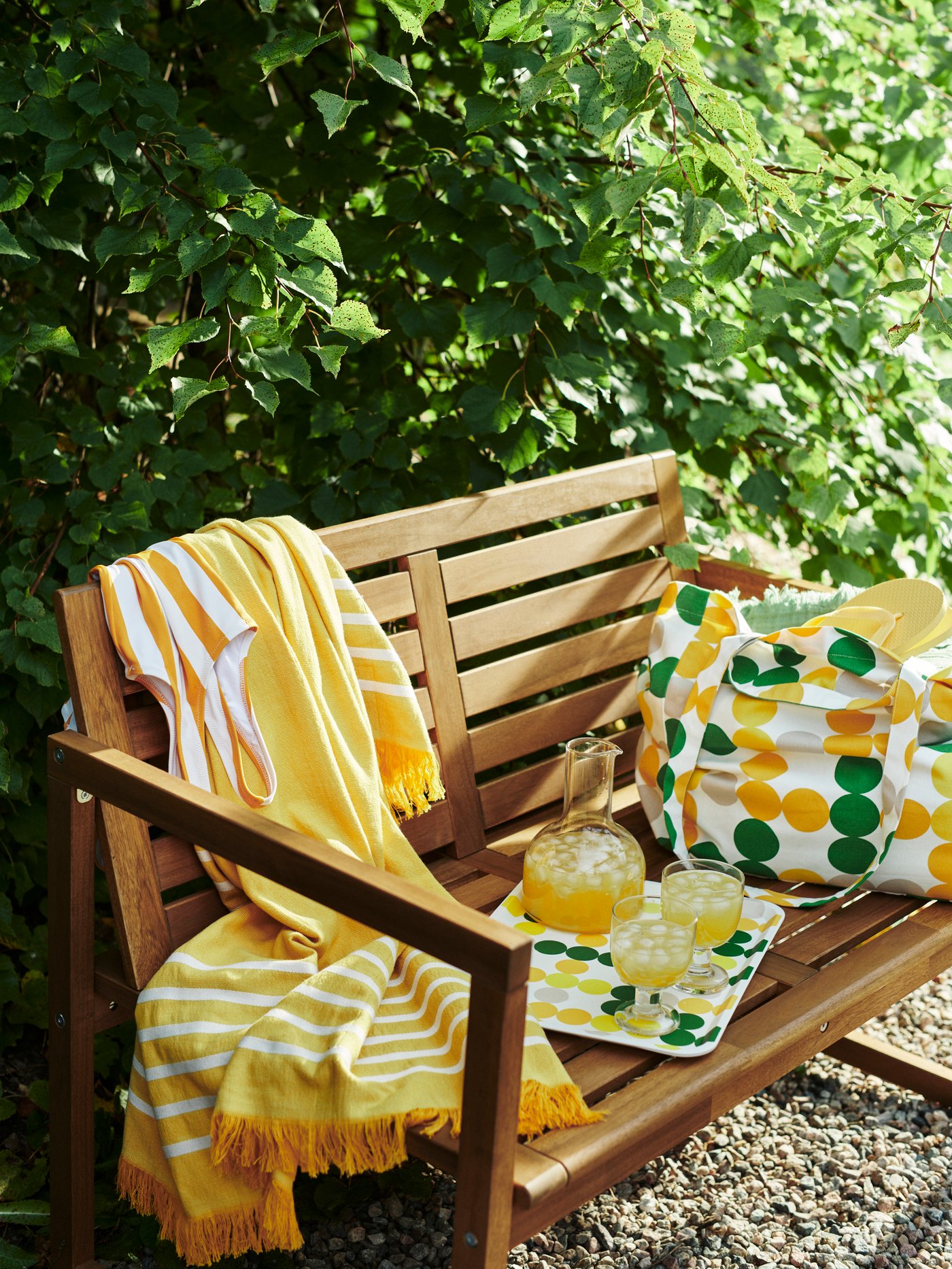 An outdoor wood bench with a colourful BRÖGGAN striped throw blanket next to a tote bag with dot pattern with a matching tray
