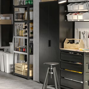 I ett garage finns svarta öppna och stängda förvaringsskåp. På hyllorna står lådor och flaskor. Under dem står återvinningskärl.