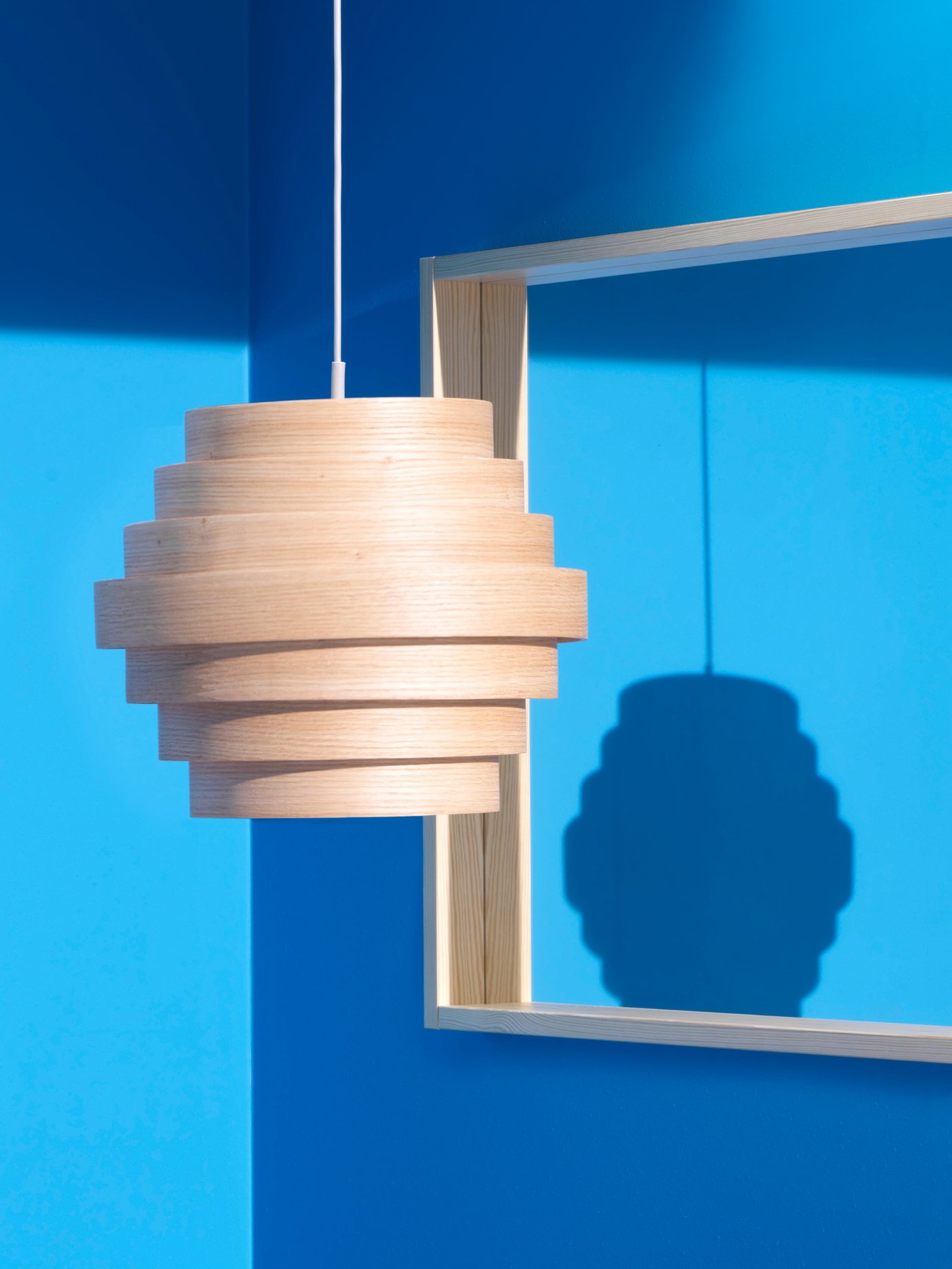 غطاء مصباح معلق DYKARKLOCKA بسبع حلقات خشبية يتدلى من سقف غرفة بحوائط باللون الأزرق الزاهي وعلى الحائط مرآة مربعة.