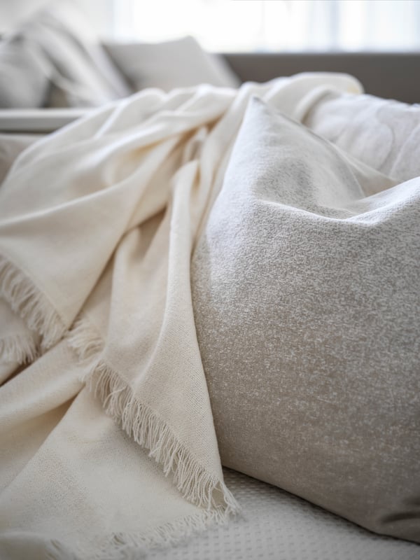 Manta INGRUN sobre uma almofada VIDESPINNARE em bege e outros têxteis em cima de uma cama individual/dupla HEMNES em branco.