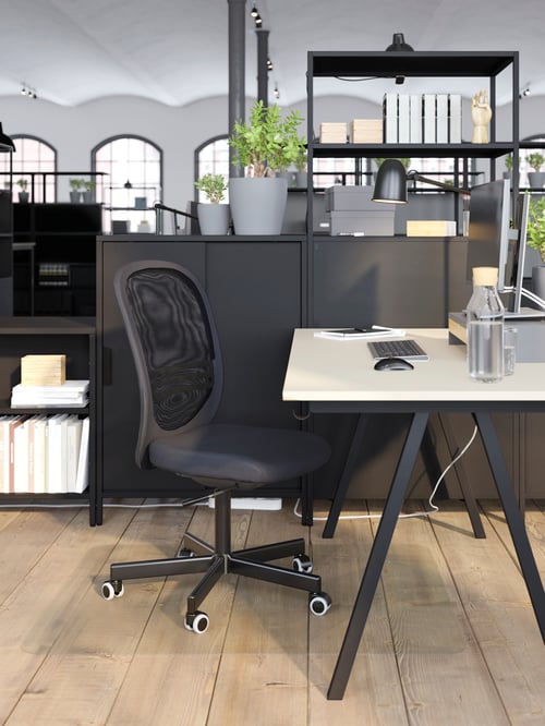 A TROTTEN desk beside cabinets and shelves with a FLINTAN chair in front of it that is on top of a clear floor protector.