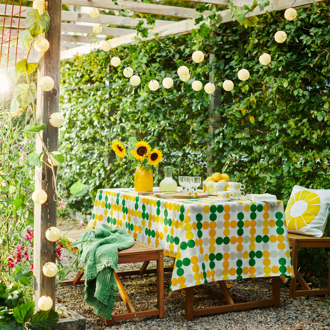 Ein gemütlich dekorierter Outdoor‑Essbereich mit einer Tischdecke in Grün‑Gelb‑Karos, umgeben von üppigem Grün. Über dem Tisch hängen warme Lichterketten, und Sonnenblumen sorgen für einen fröhlichen Frühlingsakzent.