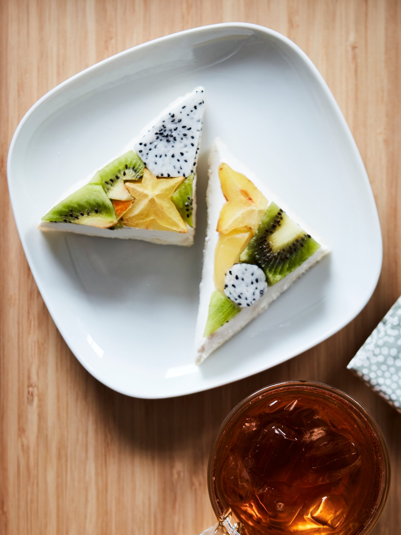 A white side plate with triangle-shaped sandwiches made with fruits like kiwi and star fruit, placed on a wooden surface.