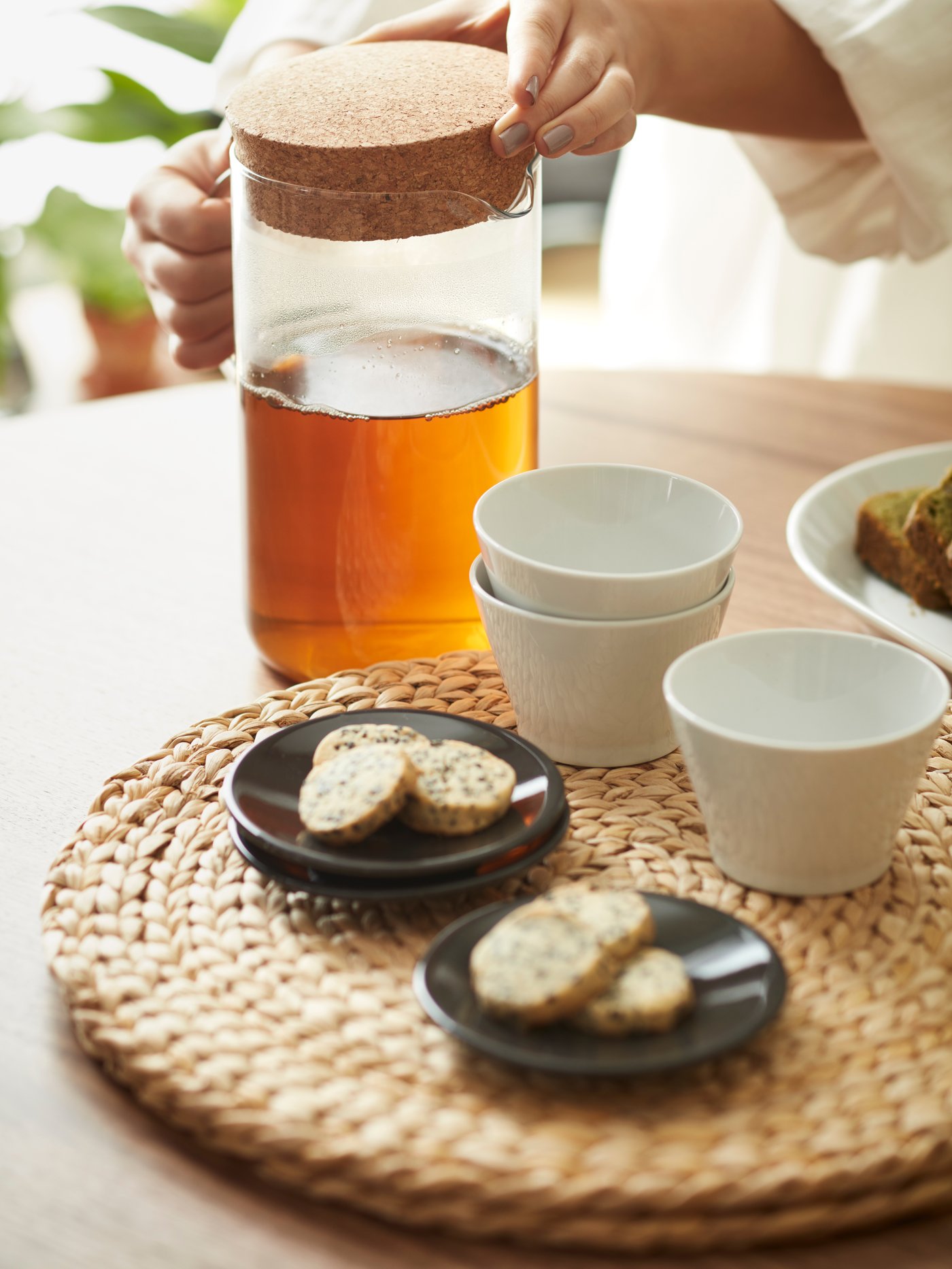 Un jarro IKEA 365+, tazas KRUSTAD de té y dos platos pequeños con galletas sobre un individual redondo SOARÉ, dispuesto sobre una mesa de madera. 