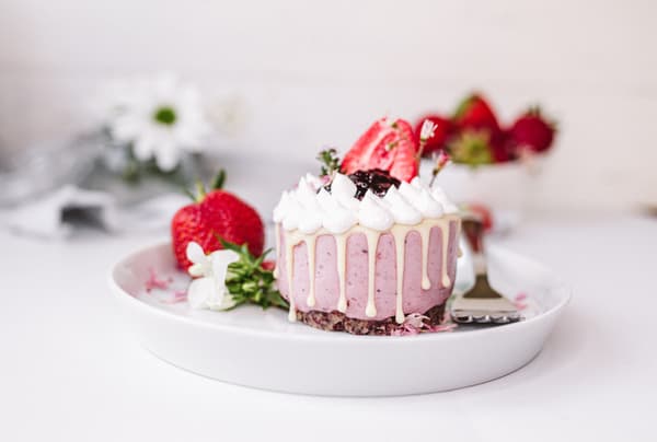 Erdbeerkuchen mit Sahne und Schokosplittern auf weißem Teller, frische Erdbeeren und Blumen im Hintergrund.