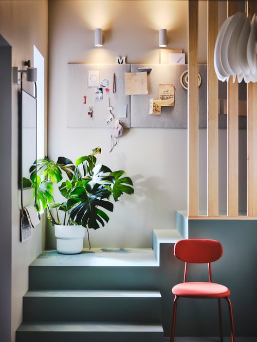 Two NYMÅNE wall downlighters cast light on memo boards which hang above a potted plant and next to a mirror and red chair.