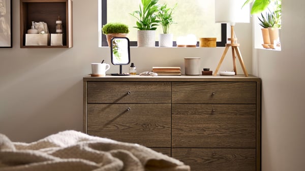 A TONSTAD chest of 6 drawers in brown stained oak veneer, a table lamp and other items on top, in the corner of a bedroom.