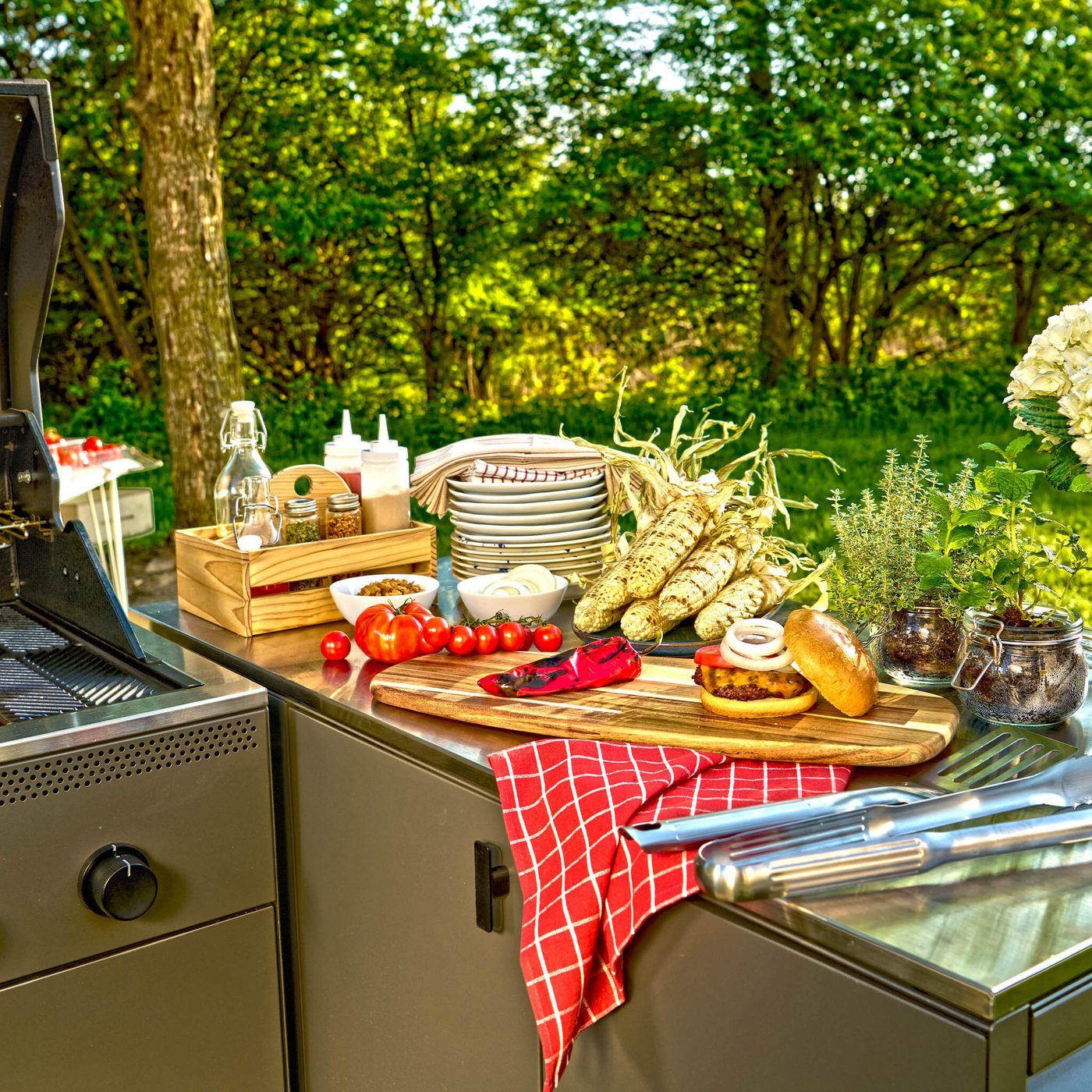Outdoor grill station with burger, vegetables, plates and herbs set up for a backyard cookout.