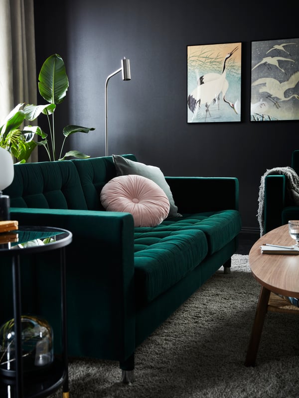 A green velvet LANDSKRONA sofa with a pink cushion on the seat and a tall plant behind it by a dark grey wall with pictures.