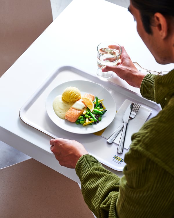 Tanji pladanj na bijelom stolu s tanjurom hrane: pečeni losos, pire krumpir, povrće (zelene mahune, grašak, mrkva) i pecivo. Uz pladanj su pribor za jelo i čaša vode.