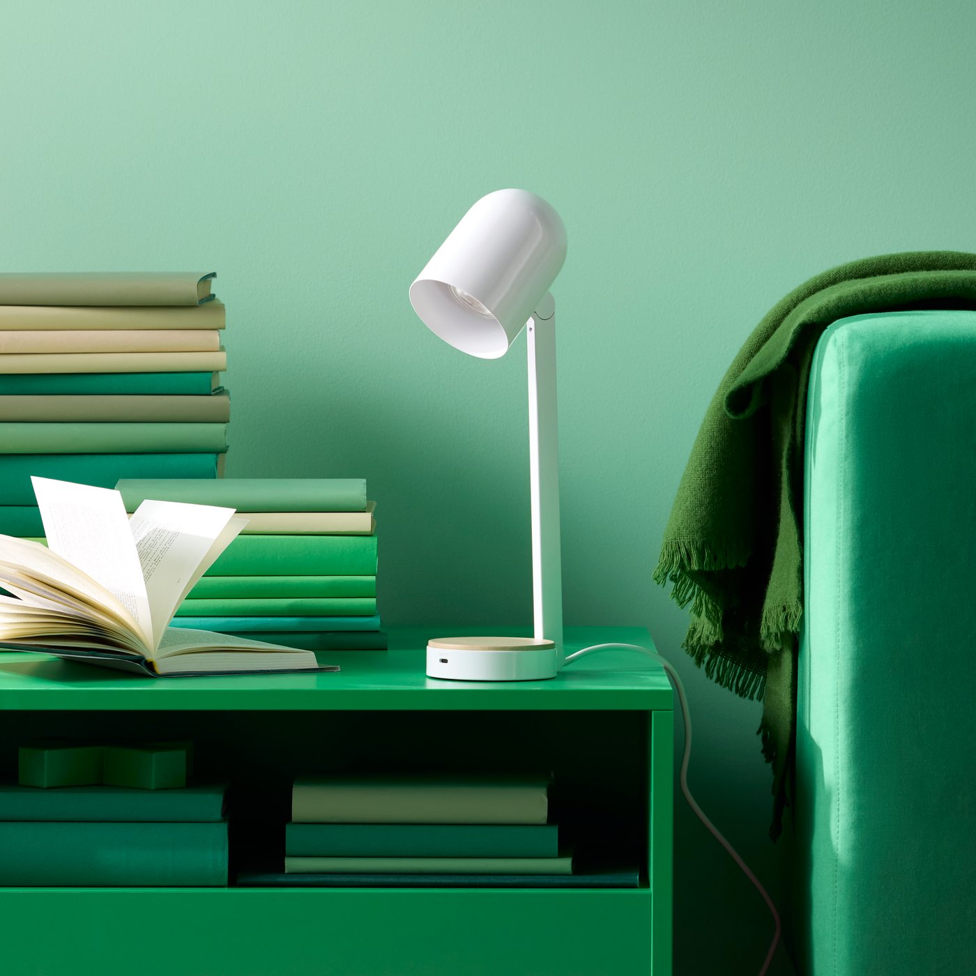 Eine weisse ENSMÄRKE Schreibtischlampe und Stapel von Büchern stehen auf einem grünen Couchtisch neben einem Sofa mit einer Decke über der Armlehne.