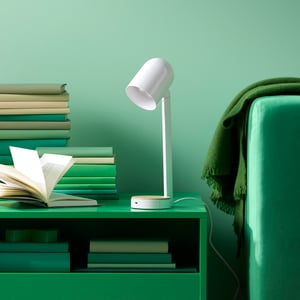 A white ENSMÄRKE desk lamp and stacks of books sit on a green coffee table next to a sofa with a throw on the armrest.