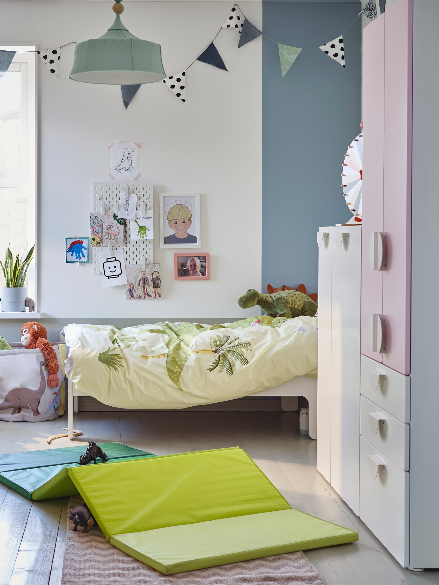 Ein Kinderzimmer mit einem Kleiderschrank in Weiß/Blassrosa, einer grünen Gymnastikmatte, einem weißen Spieltisch mit Stauraum und Stofftier-Dinosaurier.