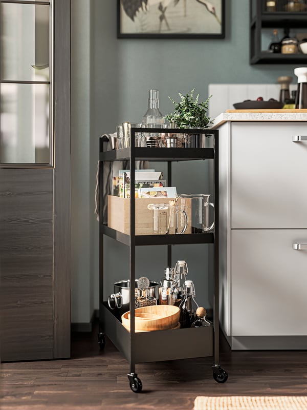 A black NISSAFORS trolley holding cookery books, glassware, bowls and more stands at the side of a row of kitchen cabinets.