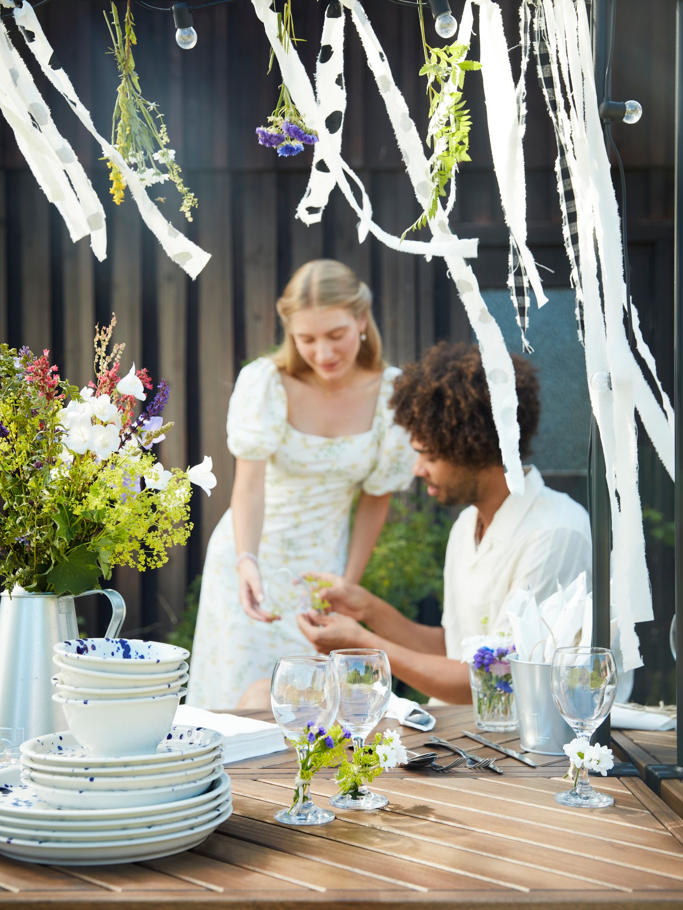 En man sitter vid ett matbord utomhus medan en kvinna står bredvid honom och arbetar tillsammans med dekorationerna till midsommarfesten.