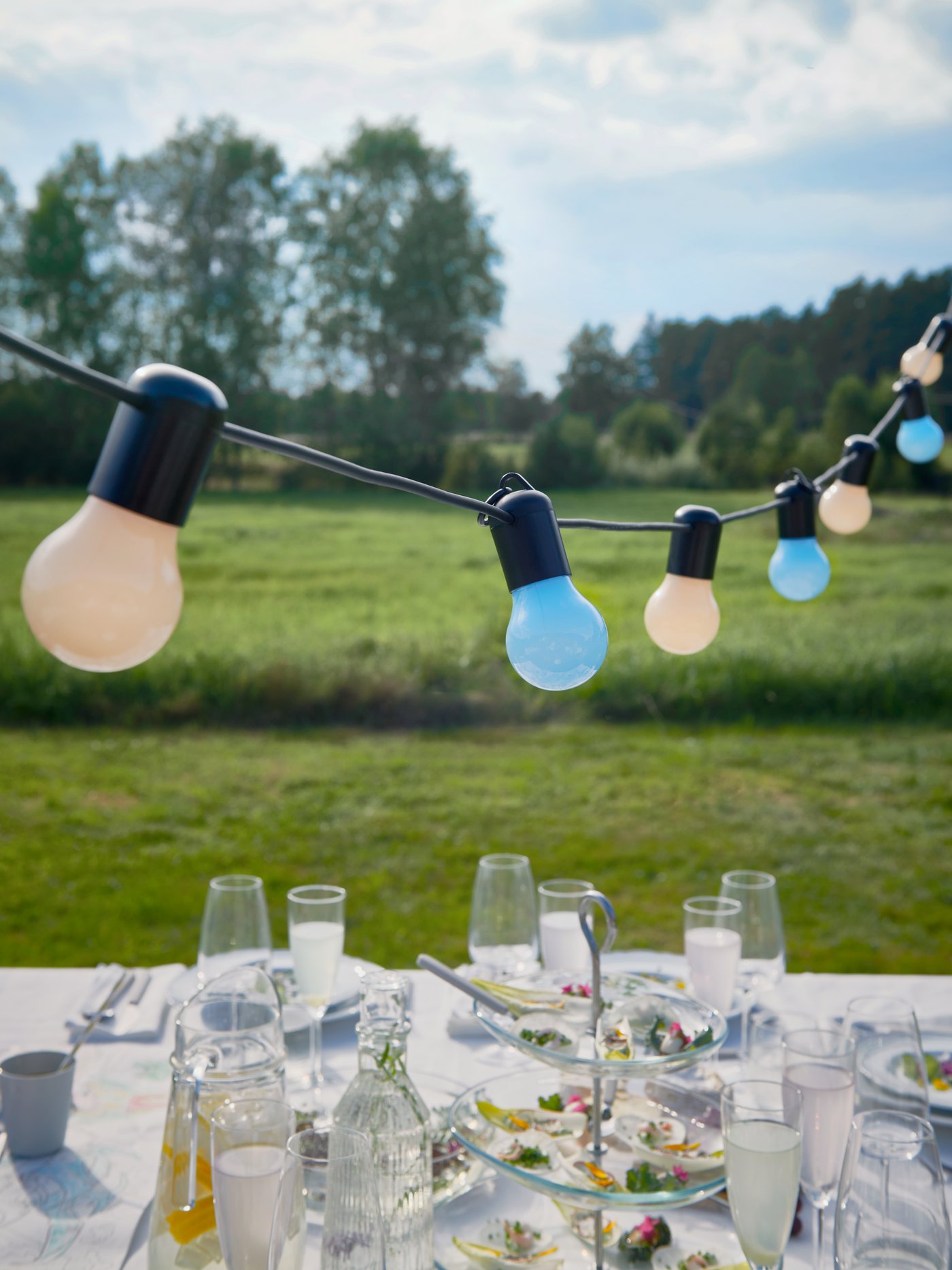 Illuminazioni a LED SOLVINDEN colorate appese sopra una tavola imbandita con cibo e bevande per una festa di matrimonio all’aperto - IKEA