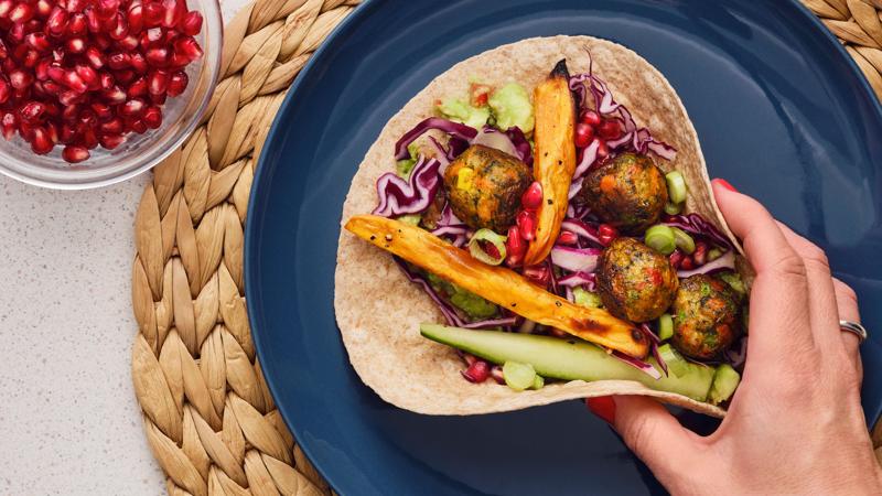 En person plockar upp en tortilla fylld med grönsaksbollar och färgglada grönsaker, placerad på en blå tallrik.