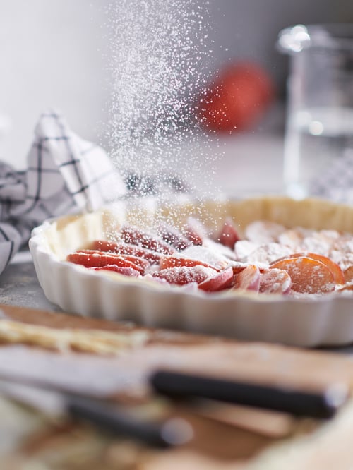 Auf einer Arbeitsplatte wird gerade Zucker über einen ungebackenen Pfirsichkuchen in einer VARDAGEN Servierform gestreut.