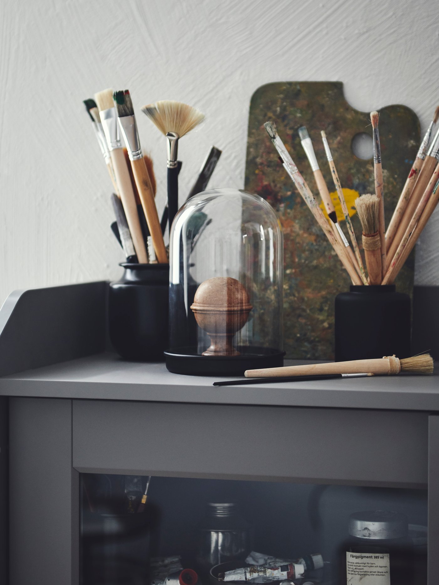 Sprawling bouquets of thin paintbrushes placed in a black KONSTFULL vase and a black jar, atop a HAUGA cabinet.