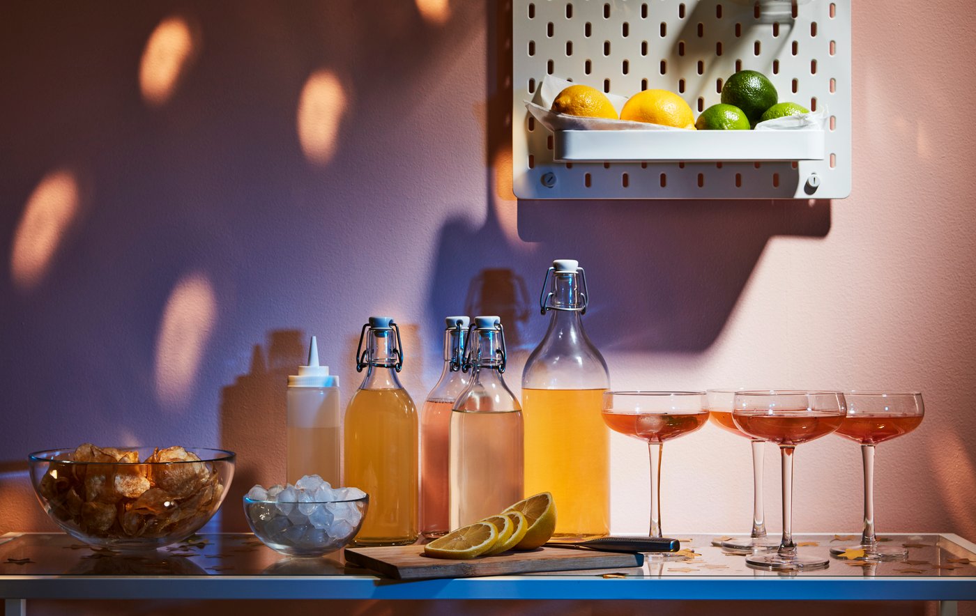 Bevande, bicchieri e accessori disposti su un tavolino. Sulla parete, un pannello portaoggetti SKÅDIS contiene limoni e lime.