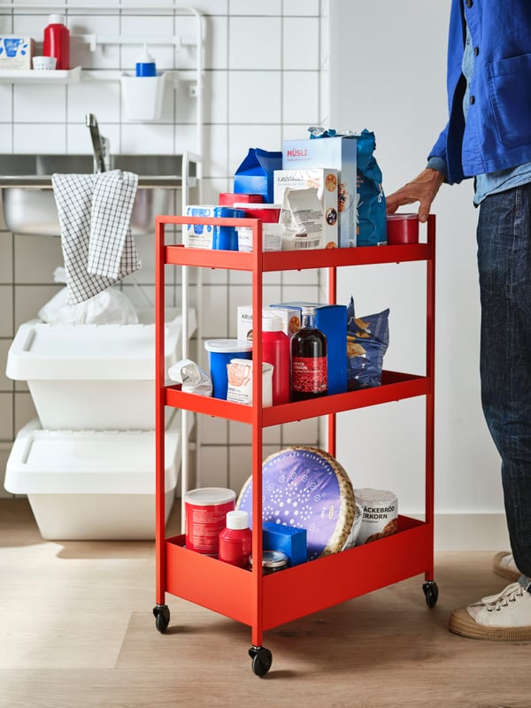 A red-orange NISSAFORS trolley filled with muesli, bread and other staples standing in front of a SUNNERSTA mini-kitchen.